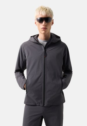 Lyshåret person iført sorte wraparound solbriller og en mørkegrå hættejakke med lynlås og hænderne i lommerne mod en ensfarvet baggrund.