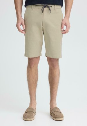 Mann trägt beigefarbene knielange Shorts mit einem Kordelzugbund und hellbraune Slipper, steht vor einem einfarbigen Hintergrund.