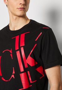 T-shirt en coton noir avec de grandes lettres et formes graphiques rouges sur le devant. Manches courtes et encolure ras du cou.