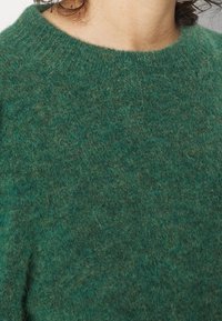 Grön ulltröja med en mjuk, fluffig textur; har en rund halsringning och en minimal design som visar en blandning av mörka och ljusa gröna nyanser.