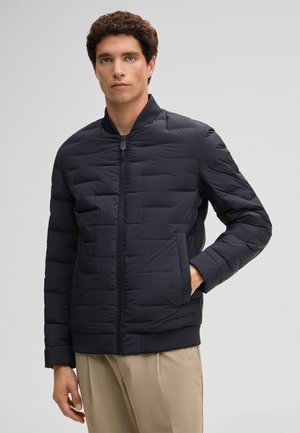 Mężczyzna ubrany w czarną pikowaną kurtkę na suwak z kołnierzem typu stójka oraz beżowe spodnie, stojący na tle gładkiego jasnoszarego tła.