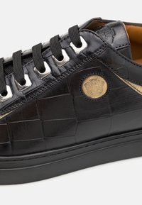 Gros plan d'une basket en cuir noir avec motif damier en relief, lacets noirs, œillets argentés et un petit emblème rond en or sur le côté.