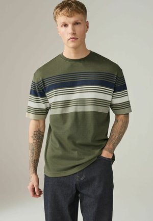 Junger Mann mit kurzen blonden Haaren und Tätowierungen an den Armen, der ein grün-navy gestreiftes T-Shirt und dunkle Jeans trägt, mit einer Hand in der Tasche stehend.