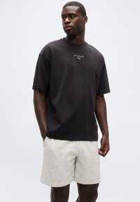 Άνδρας που φοράει μαύρο T-shirt με κοντά μανίκια και το λογότυπο "HYROX" και ανοιχτό γκρι σορτς, στέκεται με τα χέρια στις τσέπες μπροστά σε λευκό φόντο.