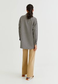 Zwart-wit pied-de-poule overhemd, oversized pasvorm, lange mouwen, met knopen afgesloten boorden; gecombineerd met lichtbeige wijde broekspijpen en sportieve schoenen.