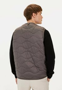 Gepolsterte graue Weste mit wellenförmigem Steppmuster, ausgestattet mit einem hohen Kragen und einem Druckknopfverschluss hinten, getragen über einem schwarzen Langarmshirt.