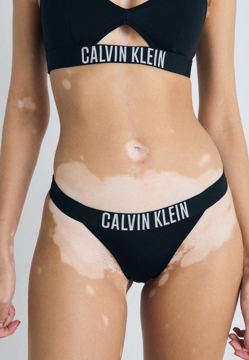 Czarny zestaw bielizny Calvin Klein składający się z braletki i stringów, z logo na pasku, wycięciem i bezszwowym wykończeniem.