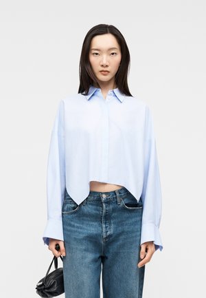 Młoda kobieta w jasnoniebieskiej krótkiej koszuli zapinanej na guziki i niebieskich dżinsach, trzymająca małą czarną torebkę na jednolitym tle.