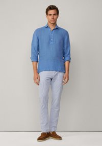 Camicia in lino azzurra chiara a maniche lunghe con un'apertura a metà bottone, abbinata a pantaloni chiari a righe e scarpe in suede marroni.