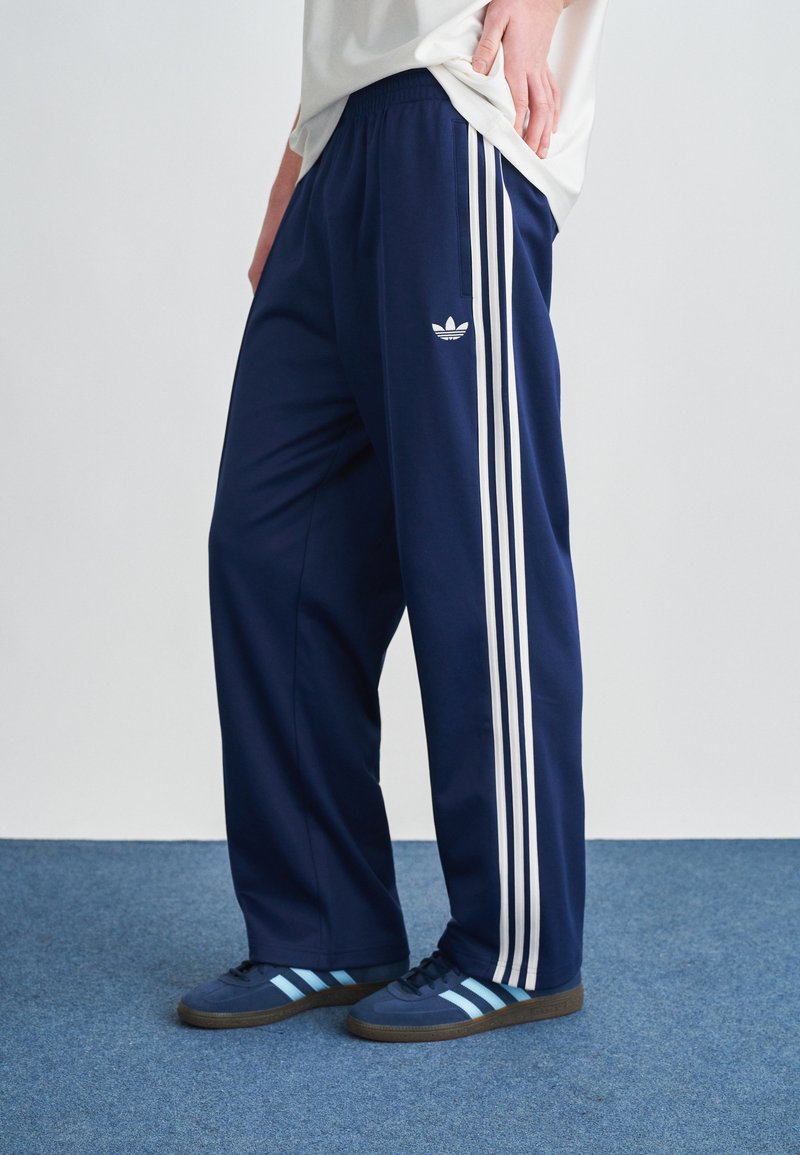 Navy plave sportske hlače s bijelim trostrukim prugama, elastičnim pojasom i malim bijelim logotipom na lijevom bedr. Imaju opušteni kroj.