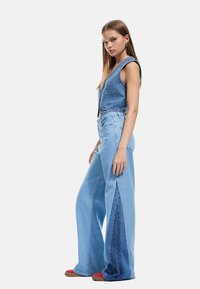 Salopette in denim blu chiaro con parte superiore senza maniche aderente, gambe ampie e pannelli laterali in blu scuro a contrasto con finitura testurizzata.