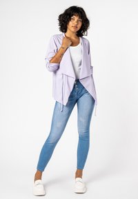Lila cardigan met lange mouwen, gedragen over een witte top. Gecombineerd met blauwe skinny jeans en witte instappers. Soepele textuur.