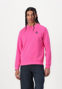 Sudadera rosa brillante hecha de tejido suave. Cuenta con cuello redondo y puños acanalados. Pequeño parche de logo negro en el lado izquierdo del pecho. Combinada con jeans oscuros.