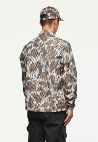 Uomo che indossa una giacca a motivo camouflage e un cappello abbinato, con pantaloni cargo neri, in piedi di schiena rispetto alla macchina fotografica su uno sfondo semplice.