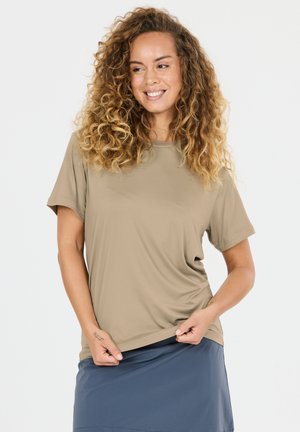Smilende kvinde med krøllet hår iført beige t-shirt og blå nederdel, der holder kanten af sin skjorte med begge hænder.