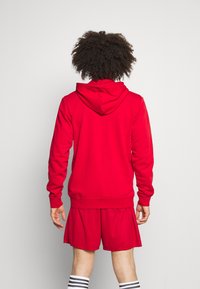 Sweat-shirt rouge avec capuche à cordon, poignets et ourlet côtelés ; porté avec un short rouge assorti. Le matériau semble être doux et flexible.