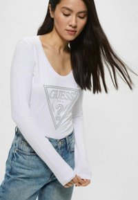 Maglietta bianca a maniche lunghe con scollatura a barca, caratterizzata da un logo grafico a triangolo argentato al centro. Abbinata a jeans azzurri chiari.
