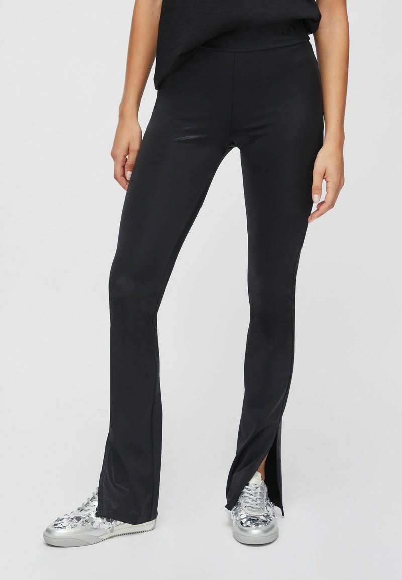 Femme portant un pantalon flare noir ajusté avec des fentes latérales, associé à des baskets argentées à sequins, debout devant un fond uni.