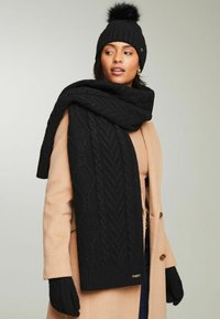 Sort strikket tørklæde med kabelmønster, parret med en sort beanie med en pom-pom og sorte handsker, alt sammen sat mod en neutral baggrund.