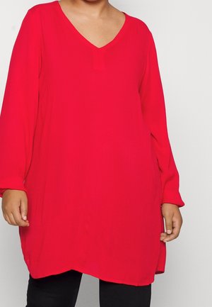 Personne portant une tunique rouge à manches longues, ample, avec un col en V, sur un pantalon noir, sur un fond uni.