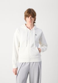 Bijela zip-up dukserica s prednjim džepom, kapuljačom s vezicom i malim zelenim logom na prsima. Napravljen od mekog materijala, ima rebraste manžete.