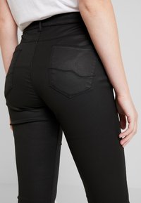 Primer plano de una persona que lleva pantalones negros ajustados con bolsillos traseros cosidos y una camisa blanca metida por dentro, ante un fondo sencillo.