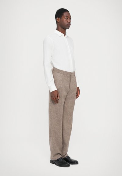 Chemise blanche à manches longues avec un col, associée à un pantalon texturé marron et des chaussures noires, montrant un profil latéral sur un fond clair.