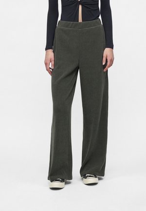 Personne portant un pantalon vert foncé à jambes larges, un haut noir à manches longues et des baskets noir et blanc, debout devant un fond uni.