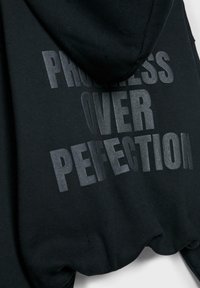 Črna jopica s kapuco z izbočenim temno sivim napisom "NAPREDEK NAD POPOLNOSTJO" na hrbtni strani, prikazana obešena na belem ozadju.