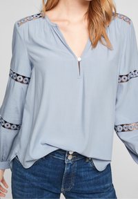 Vaaleansininen pusero V-päällä, pitkät hihat, joissa on pyöreitä pitsikoristeita, ja nappi kauluksessa. Tasainen kangas, rento malli.