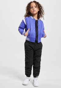 Blå varsity-jacka med vita ränder, svarta cargobyxor och vita sneakers. Jackan har knappar fram och fickor.