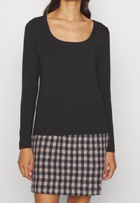 Kvinna som bär en svart långärmad topp med rund halsringning tillsammans med en knälång beige och svart rutmönstrad kjol.