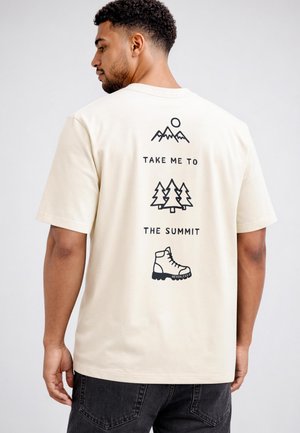 Mann mit lockigem Haar, der ein cremefarbenes T-Shirt mit schwarzen Linienzeichnungen von Bergen, Bäumen, Wanderschuh und dem Text „Bring mich zum Gipfel.“ trägt.
