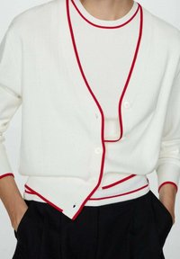 Cardigan bianco a coste con rifiniture rosse e bottoni, indossato sopra una maglietta bianca. Presenta maniche lunghe e una vestibilità comoda. Accentuato da un dettaglio del logo.