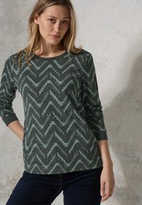 Haut à manches longues en vert foncé avec un motif en zigzag bleu clair. Encolure ronde et tissu texturé, avec une coupe décontractée.