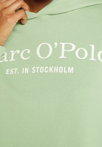 Sudadera verde menta de tela suave, con un bolsillo frontal, capucha con cordón y gran texto blanco: "Marc O'Polo EST. EN ESTOCOLMO."