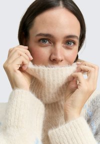 Pull en maille écru à col roulé avec une finition texturée et duveteuse ; le modèle relève le col ; tissu doux avec des bords roulés.