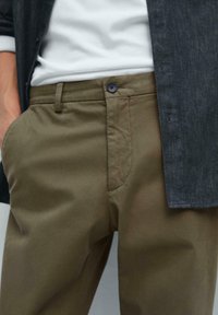 Pantalons vert olive avec une texture lisse, poches avant et fermeture à bouton unique. Associé à une chemise à manches longues noire.