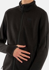 Chaqueta de forro polar negra con cremallera, que cuenta con un alto cuello, tejido texturizado y bolsillos laterales con cremallera. Incluye agujeros para los pulgares en los puños para mayor comodidad.