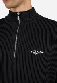 Black ribbed sweater with high collar, half zipper, and white embroidered logo; textured knit fabric provides a cozy appearance.