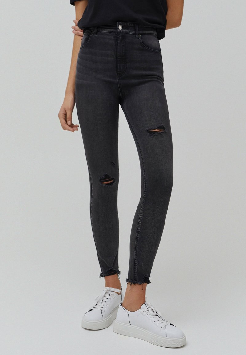 Mustat korkeavyötäröiset kapeat farkut, joissa on kulutettuja yksityiskohtia, repsottavat reunat ja sileä farkkukangas, yhdistettynä valkoisiin lenkkareihin.