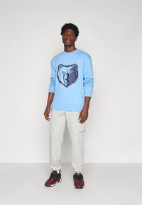 Világoskék pulóver, amelyen egy medve grafika található, szürke cargo melegítőnadrággal és fekete edzőcipővel, piros kiegészítőkkel.