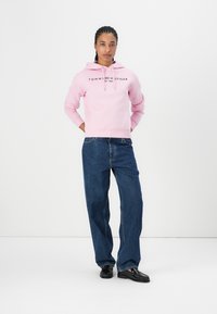 Ružová mikina s logom "TOMMY HILFIGER", v kombinácii s rovnými modrými džínsami a čiernymi topánkami. Hladká textúra, neformálny dizajn.