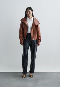 Žena u smeškastoj kožnoj jakni, bijeloj košulji s dugmetima, crnim kožnim hlačama i zlatnim cipelama s oštrim vrhom, stoji naslonjena na jednostavnu sivu pozadinu.