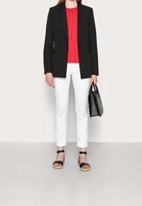 Blazer negro sobre una blusa roja fruncida, pantalones tobilleros blancos y sandalias negras. Sosteniendo un pequeño bolso negro. Texturas suaves, ajuste a medida.