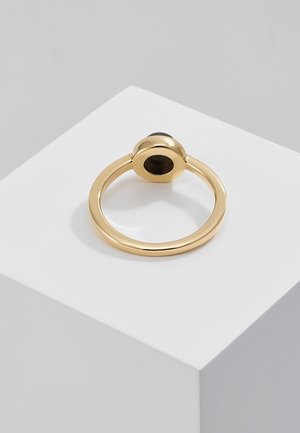 Złoty pierścionek z gładką, okrągłą obręczą, na której osadzony jest pojedynczy czarny owalny kamień w podniesionej oprawie, prezentowany na białym stojaku.