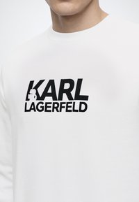 Λευκό πουκάμισο με έντονο μαύρο κείμενο "KARL LAGERFELD" και μια μικρή σιλουέτα ενός ατόμου με γυαλιά ηλίου και αλογοουρά ενσωματωμένη στο γράμμα Κ.