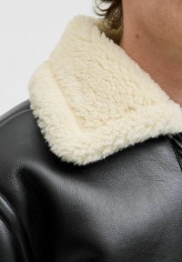 Nærbillede af en sort læderjakke med en tyk, crèmefarvet shearling-krave, båret af en person.