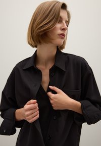 Camicia nera, oversize, con bottoni in morbido tessuto, colletto e taschino sul petto; il modello regola la camicia all'uscita del collo.