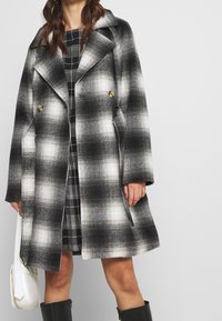 Cappotto a quadri bianco e nero con colletto oversize, due bottoni e tasche laterali. Indossato sopra un vestito a quadretti bianchi e neri. Tessuto liscio.